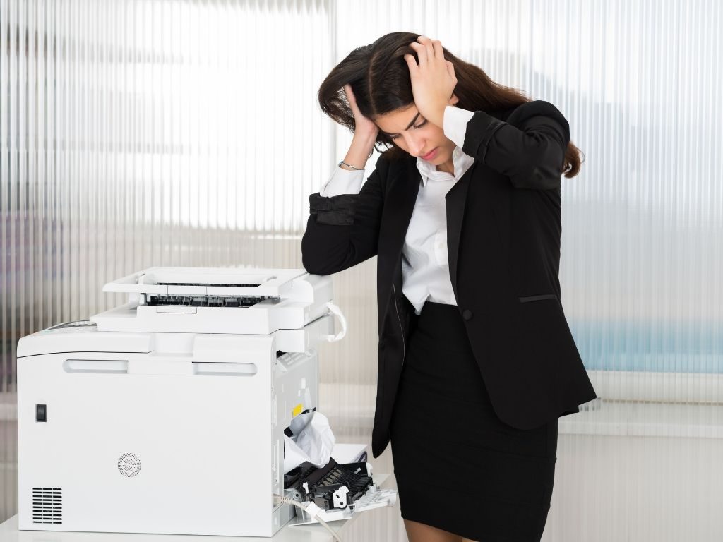 A woman standing next to a printer holder her head in frustration.
